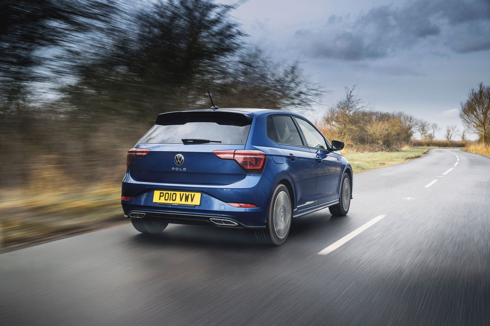 A blue Volkswagen Polo drives on a country road with blurred motion, under a cloudy sky, surrounded by bare trees.