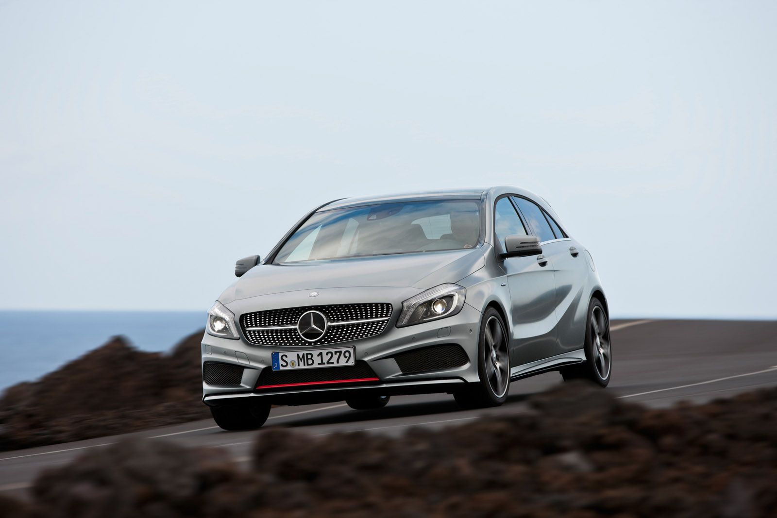 Silver Mercedes-Benz car driving on a coastal road with a clear sky and rocky foreground.