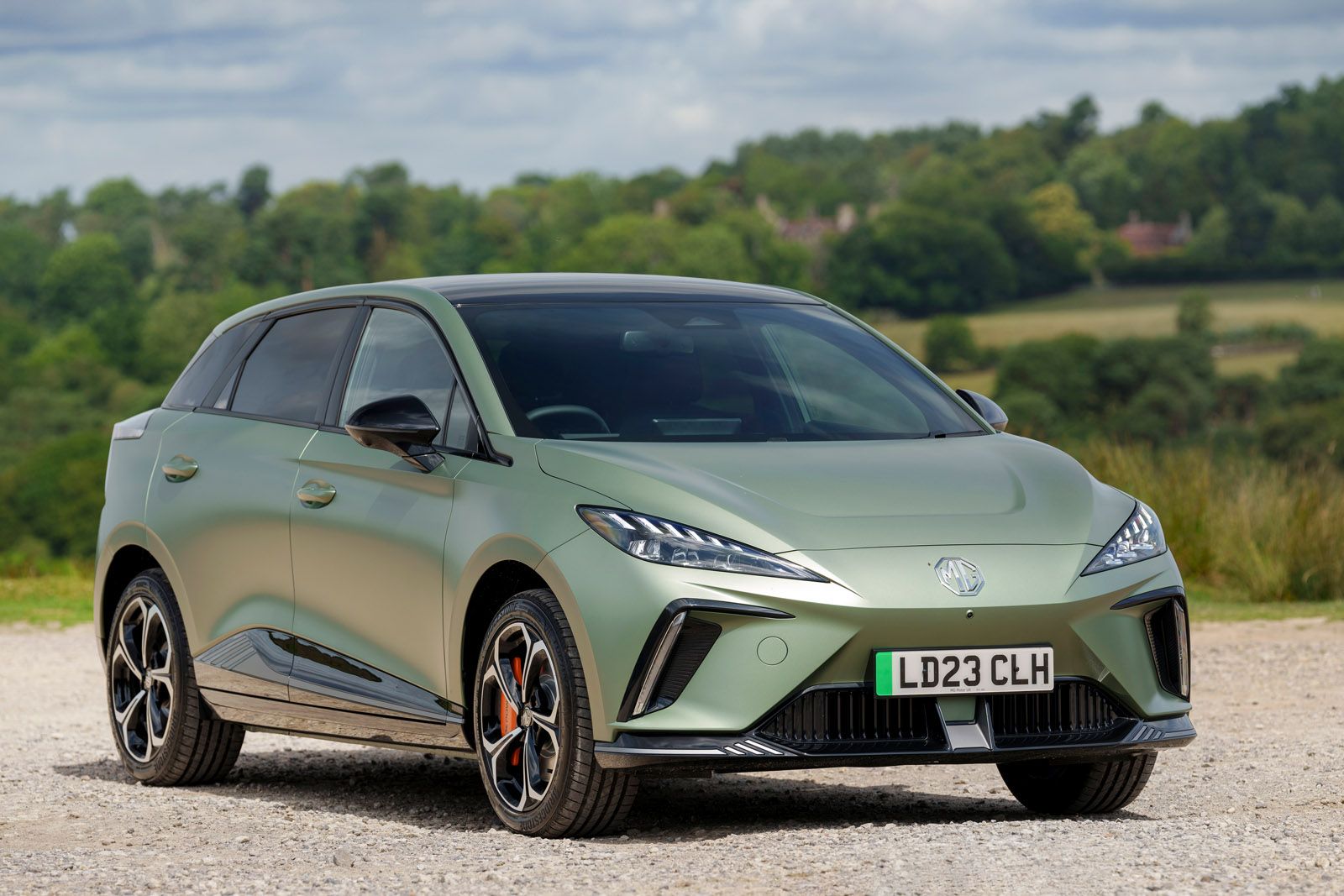 A green, modern hatchback parked on a gravel path with a scenic, wooded background under a cloudy sky.
