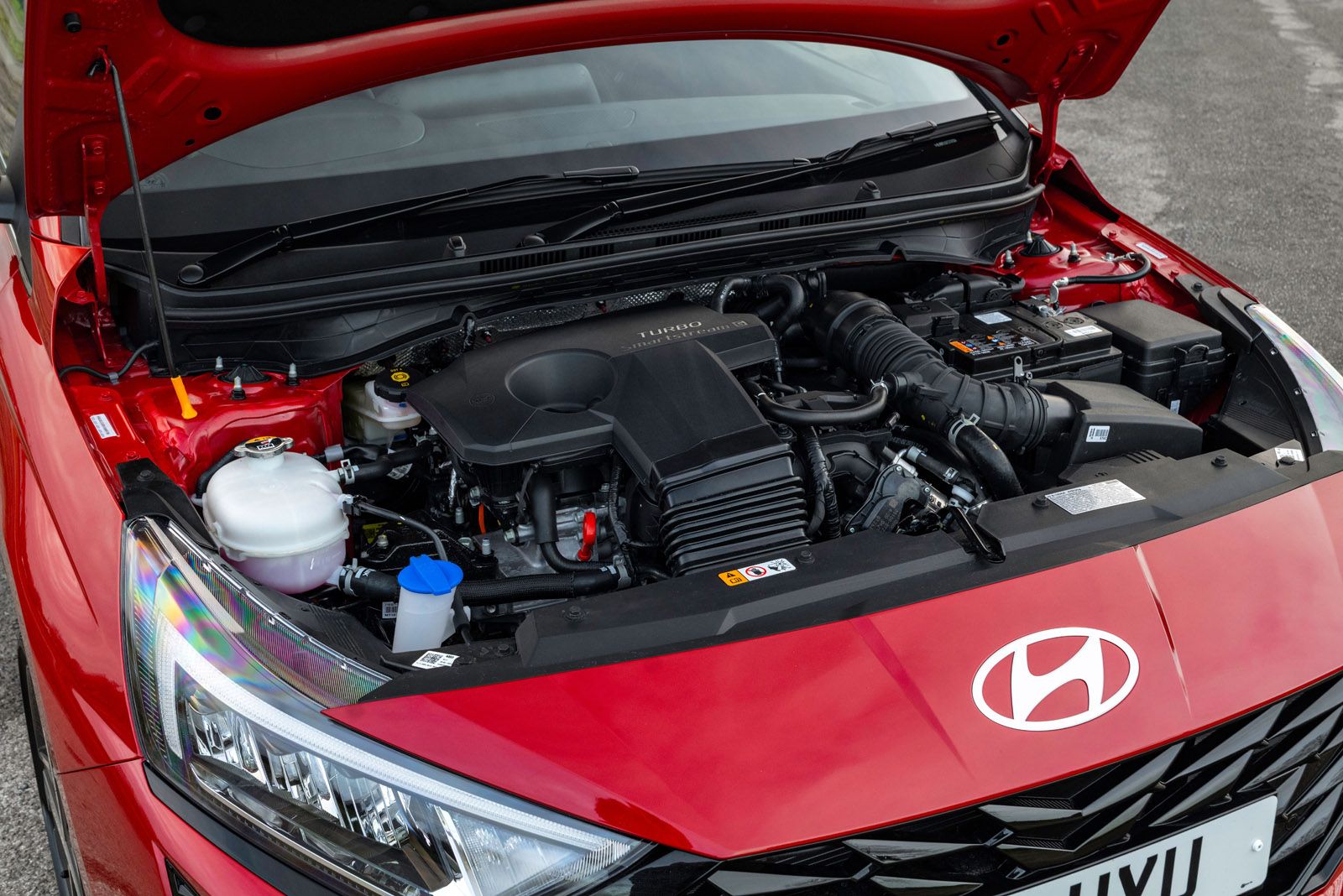Red Hyundai car with an open hood showing the engine compartment, including various components and fluid reservoirs, parked on a paved surface.