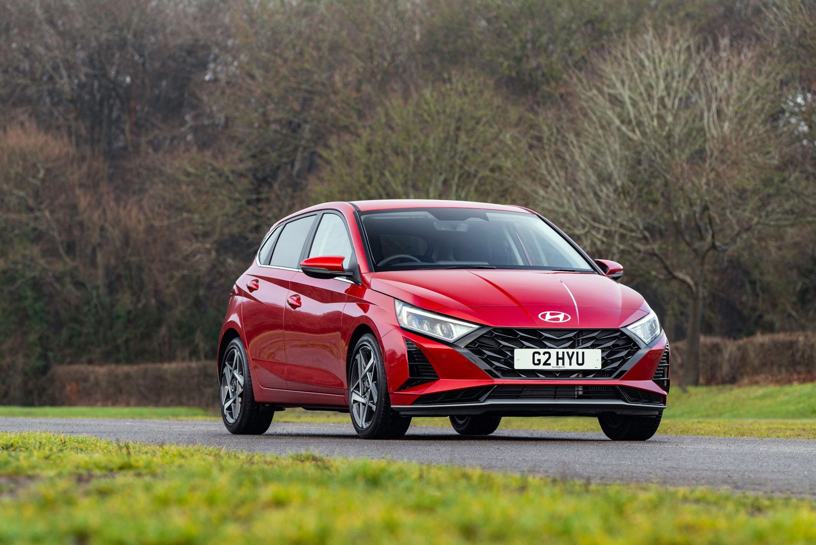 A red Hyundai hatchback parked on a rural road, with bare trees and grass in the background under an overcast sky.