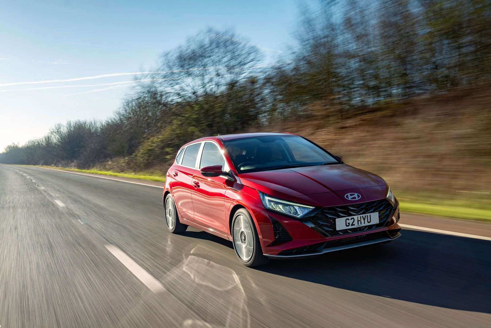 Red Hyundai hatchback driving on a sunny road with blurred trees in the background.