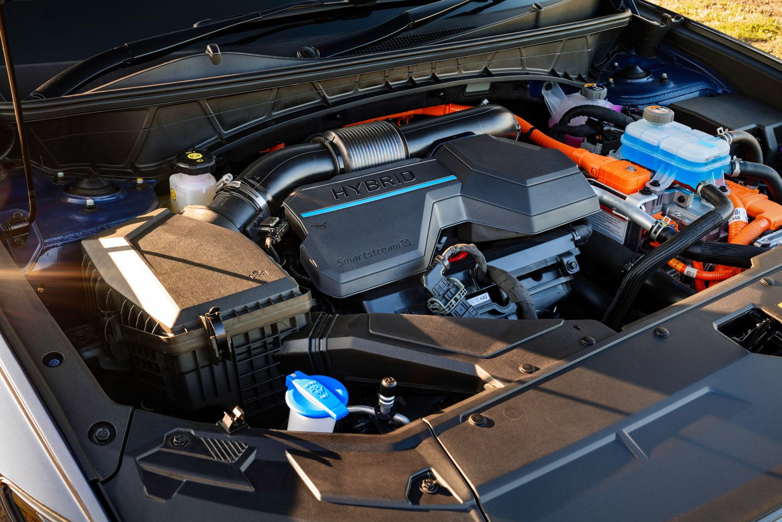 Close-up of a car engine labeled "HYBRID." It features various components, including hoses, wiring, and fluid containers, with sunlight illuminating part.