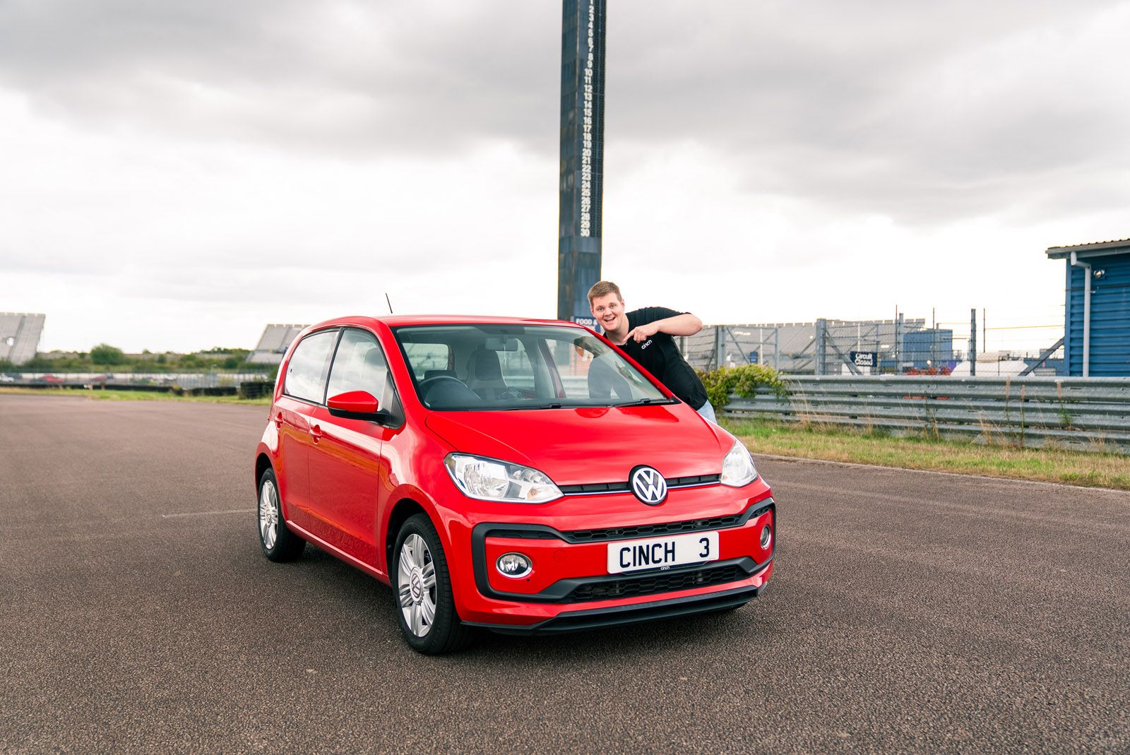 A person leans on a red Volkswagen parked on a race track, with a tall scoreboard in the background under a cloudy sky.