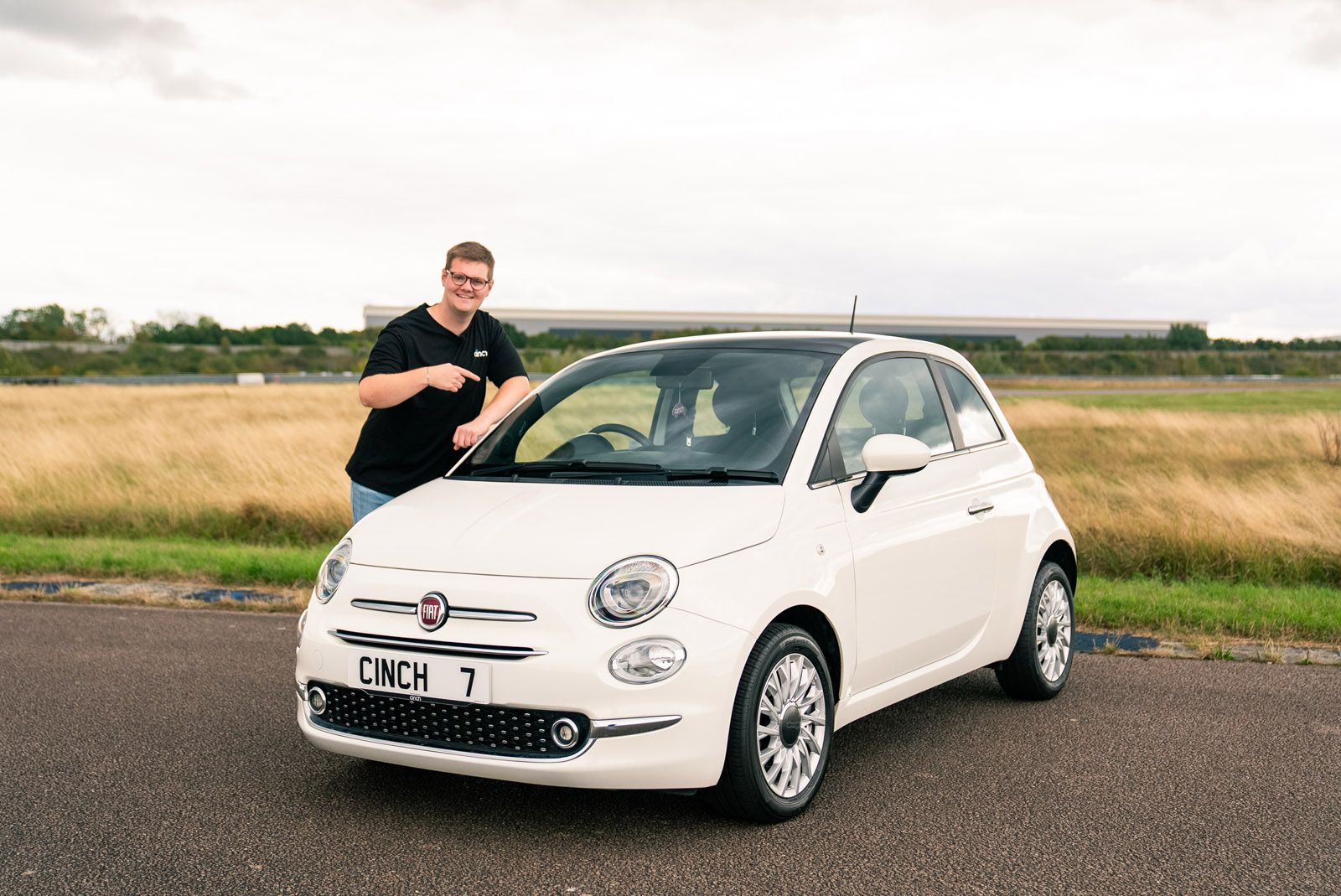 A person standing next to a white Fiat 500 with a "CINCH 7" license plate, smiling, in a grassy outdoor setting.