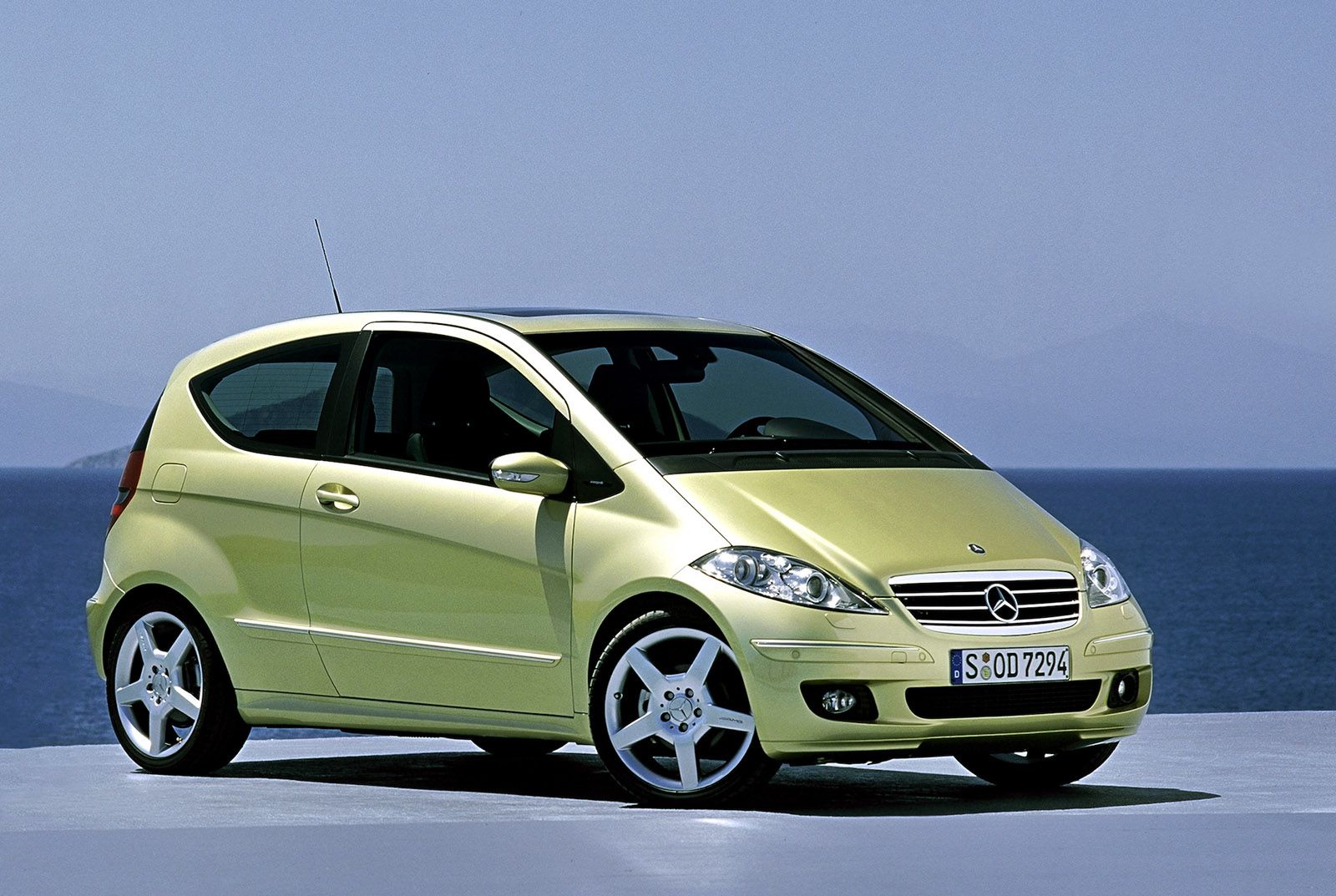 A compact yellow car parked on a coastal road with a clear blue sky and ocean in the background. Front and side view of the vehicle.