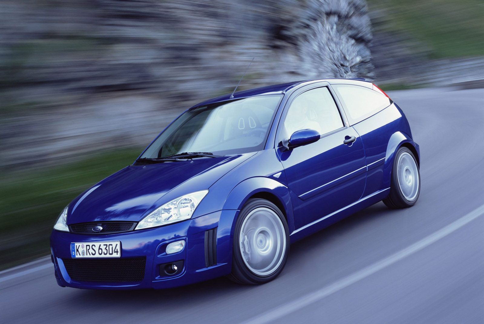 A blue Ford hatchback speeds along a curving road, with blurred motion background suggesting fast movement.
