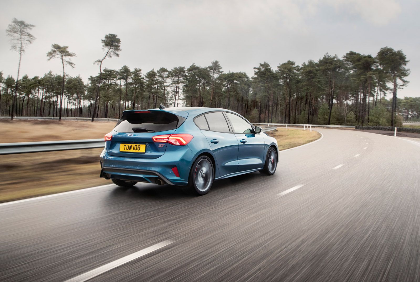 A blue car with a license plate "TUN 108" drives on a curved road surrounded by trees under a cloudy sky.