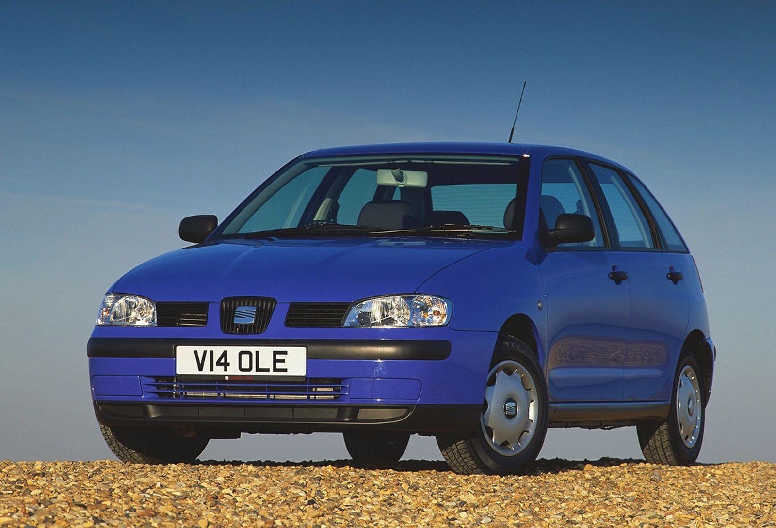 Blue compact car parked on a gravel surface, featuring a license plate with "V14 OLE," set against a clear blue sky.