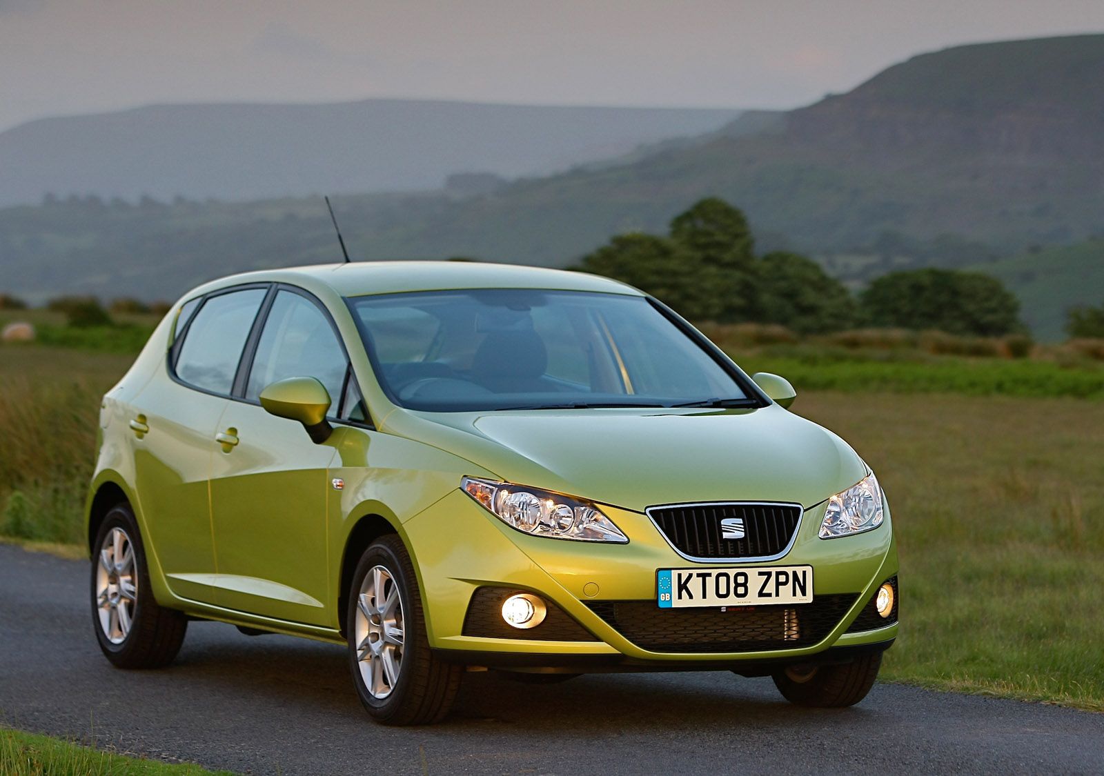 Green hatchback car parked on a rural road, with hilly landscape and soft evening light in the background.