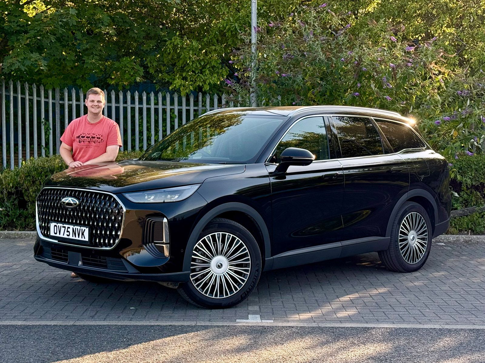 A person in a red shirt stands beside a sleek, black SUV parked on a street, with greenery and a fence in the background.