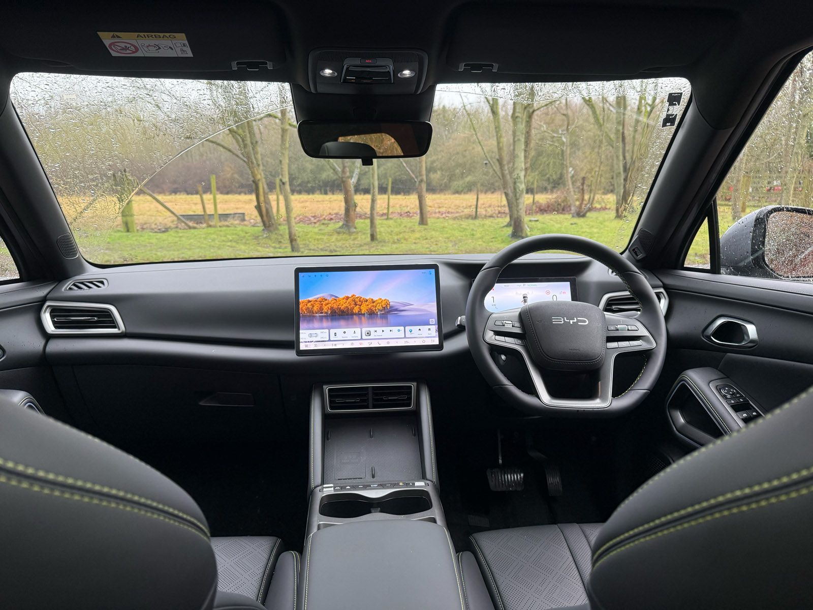 Car interior with a modern dashboard, dual screens, and steering wheel, viewed from the back seat. Rainy landscape visible through the windshield.