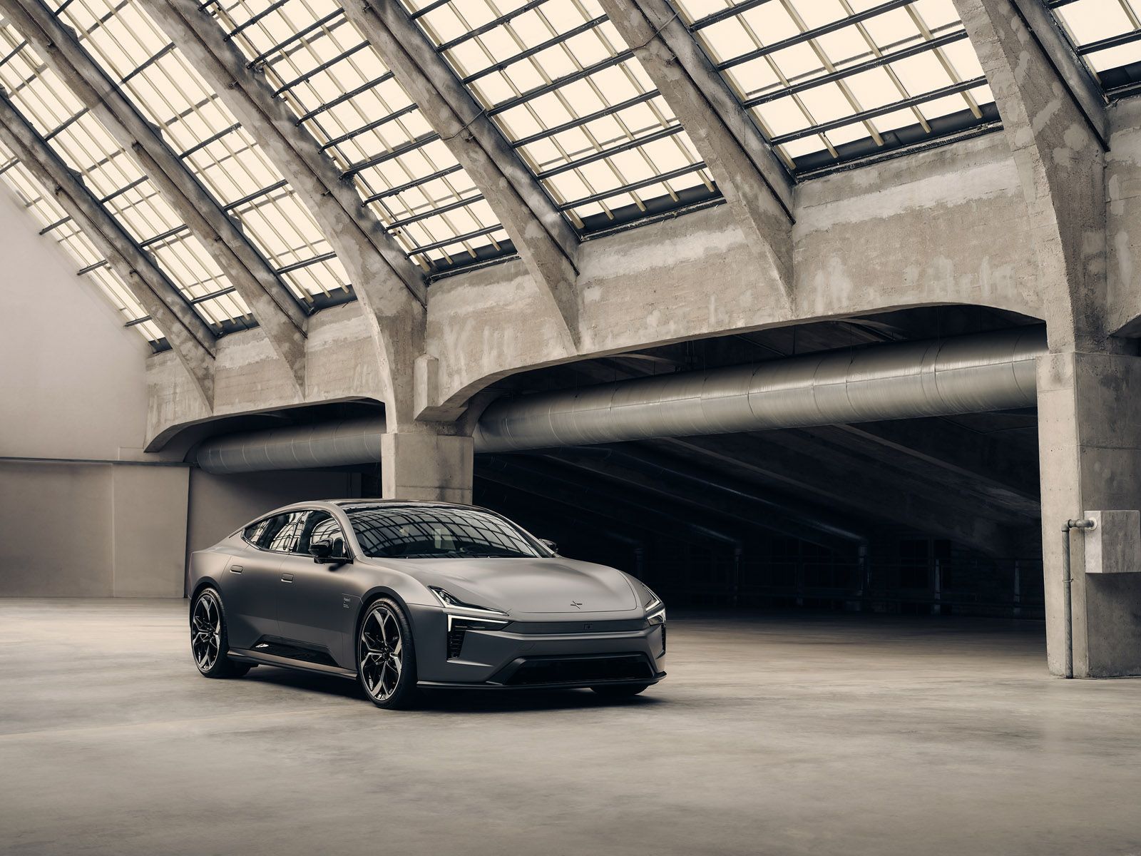 A sleek, grey Polestar 5 sports car parked inside an industrial building with large windows and concrete beams.