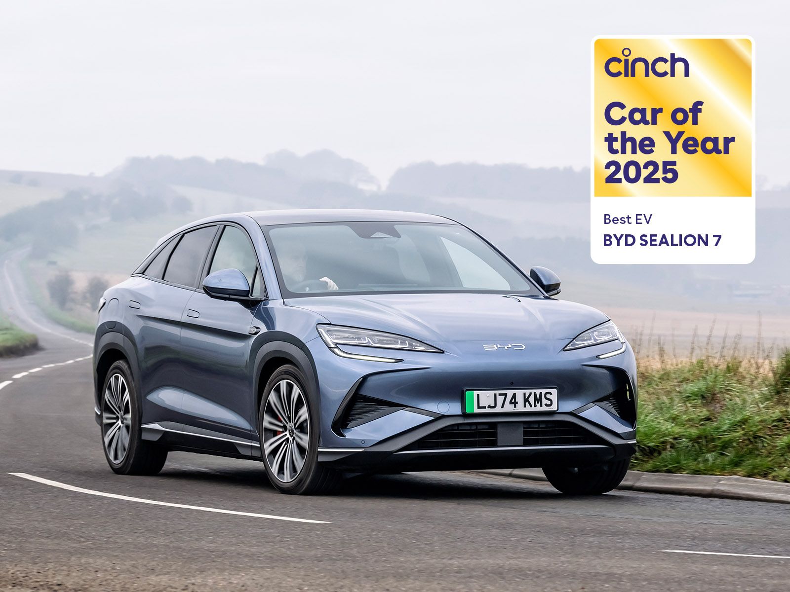 Blue BYD Sealion 7 driving on a scenic road, awarded "Car of the Year 2025" and "Best EV" by cinch, with a sign in the top right corner.