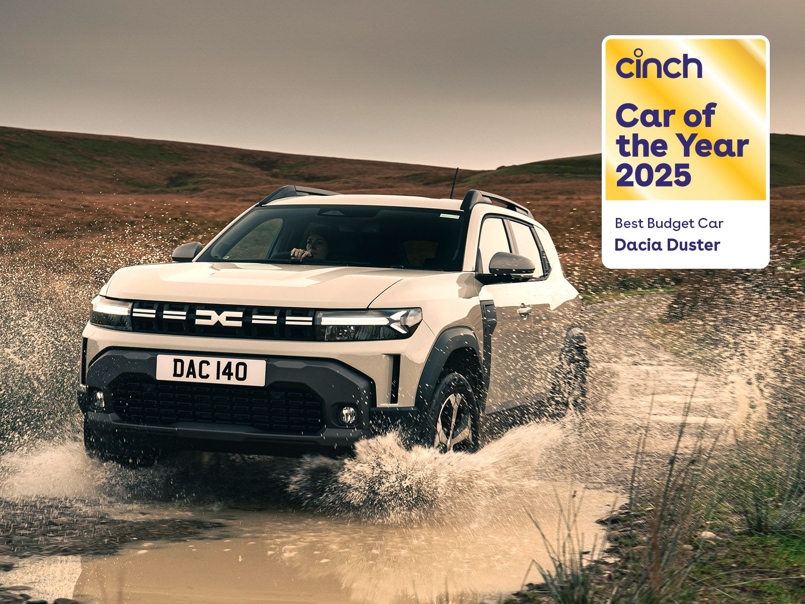 A white Dacia Duster drives through a muddy puddle in a rural landscape. A badge reads "cinch Car of the Year 2025, Best Budget Car."