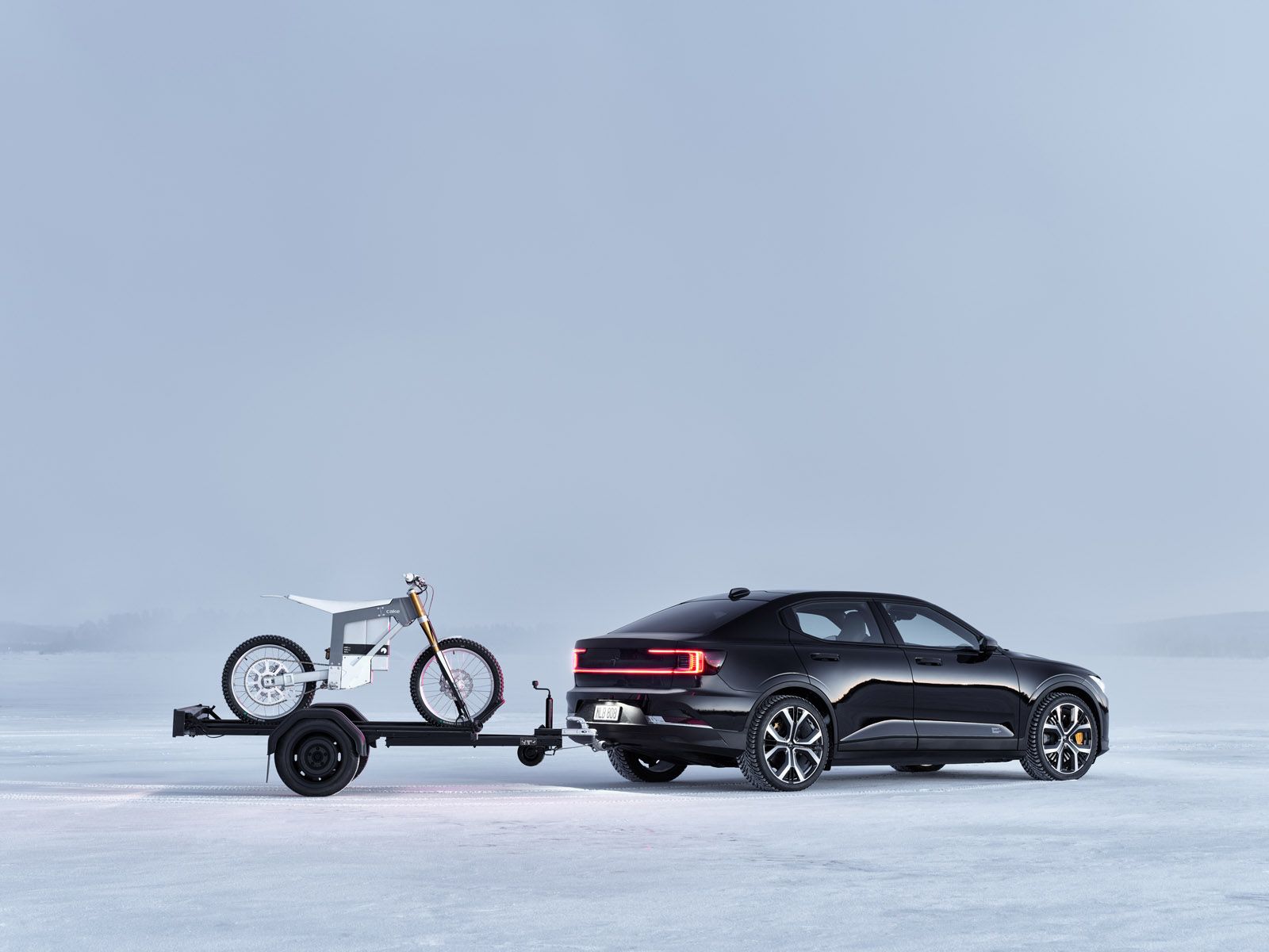 A black car towing a trailer with a white electric motorcycle on a snowy landscape under a cloudy sky.