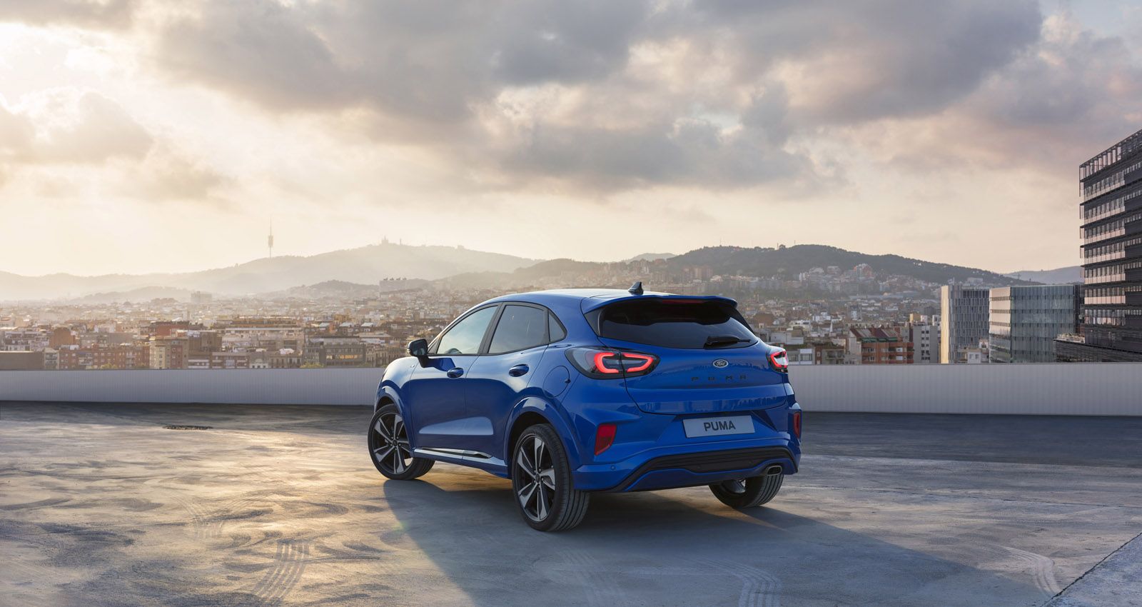 A blue Ford Puma SUV is parked on a rooftop with a cityscape and mountains in the background under a cloudy sky.