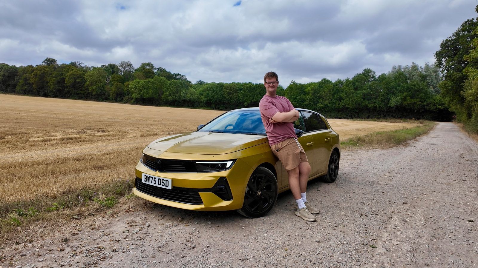 Person in pink shirt leans on a gold car parked on a rural road, with fields and trees in the background under a cloudy sky.