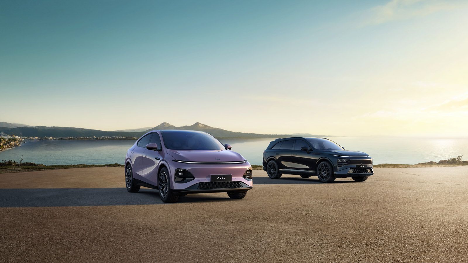 Two sleek electric SUVs parked on a coastal road, with a scenic ocean and mountain backdrop under a clear sky.