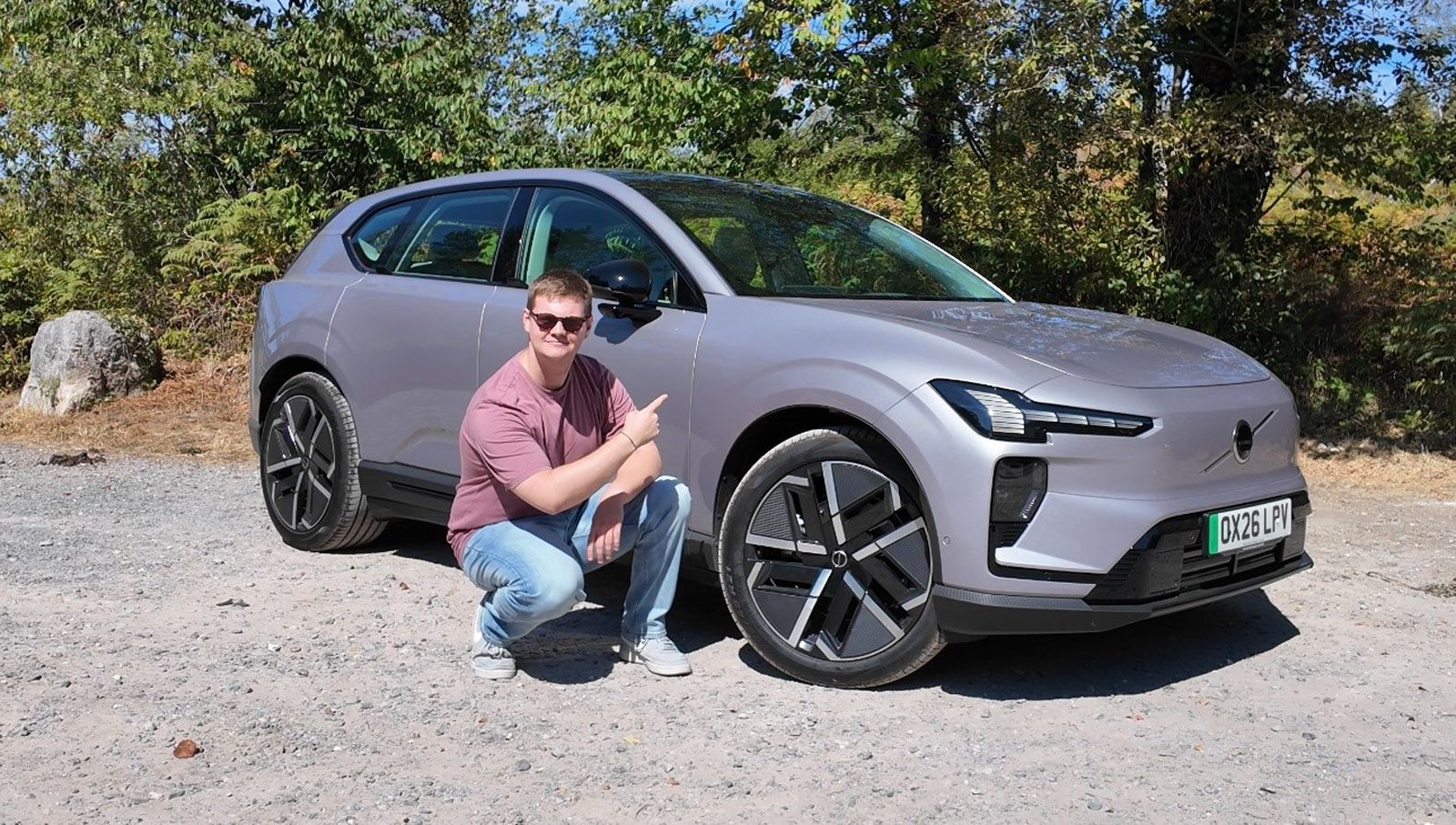 Person smiling and pointing at a modern silver car with unique wheel design, parked on a gravel path with trees in the background.