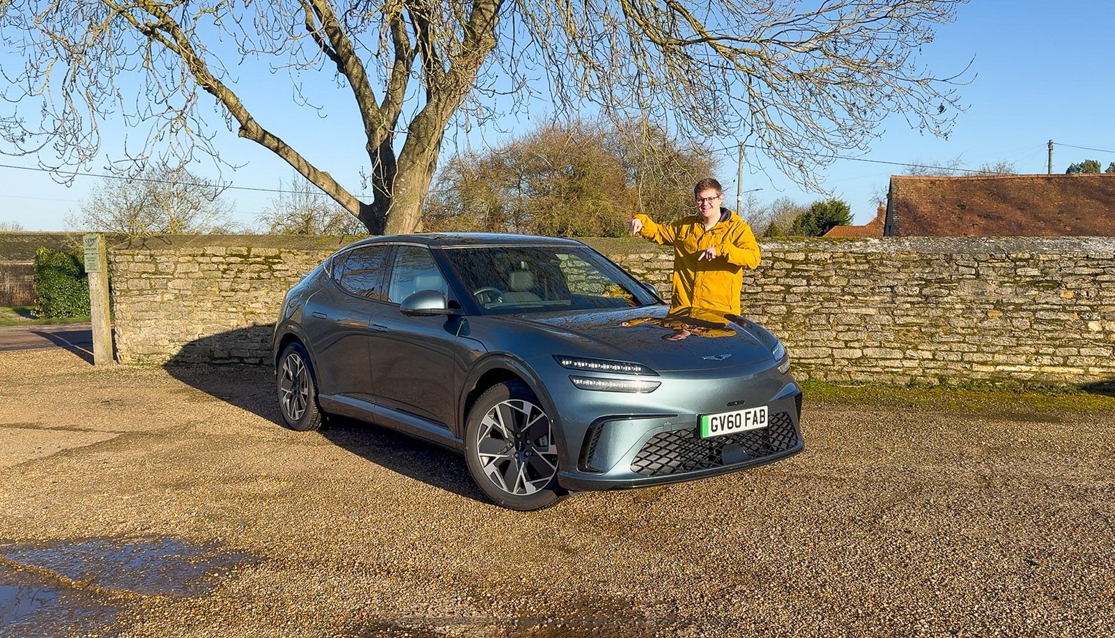 Person in a yellow jacket giving a thumbs up next to a parked gray sedan on a sunny day, with a stone wall and trees in the background.