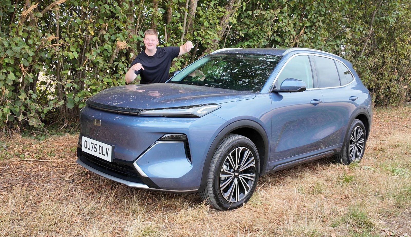 A person stands next to a blue SUV in a grassy area, smiling and gesturing. The car's license plate reads "OU75 DLV."