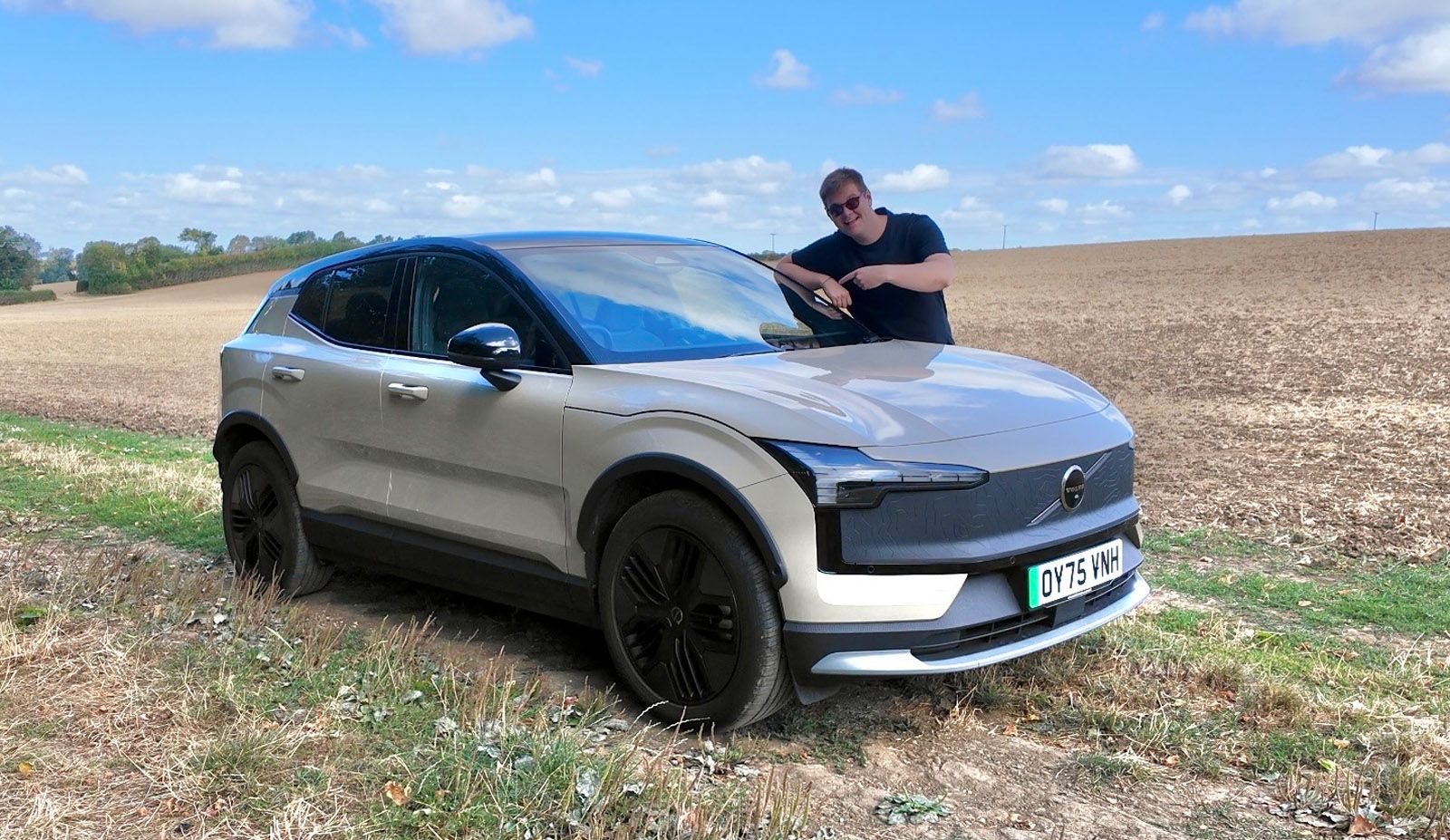 A person leans on a silver SUV with black accents, parked on a grassy field under a blue sky with scattered clouds.