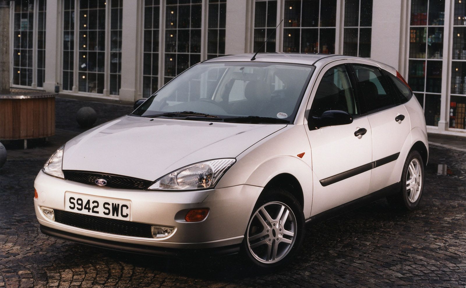 Silver Ford hatchback parked on a cobblestone street in front of a building with large windows.