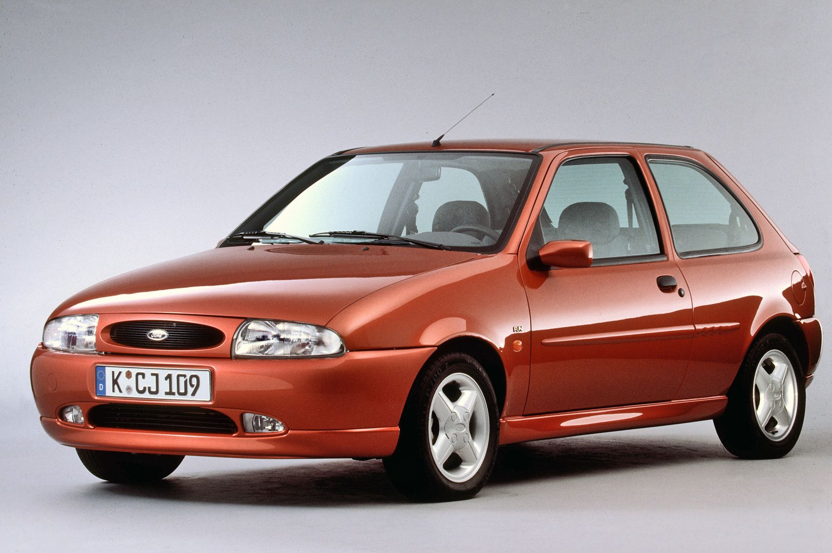 A red, three-door hatchback car with alloy wheels is parked on a plain background. The vehicle has a European license plate.