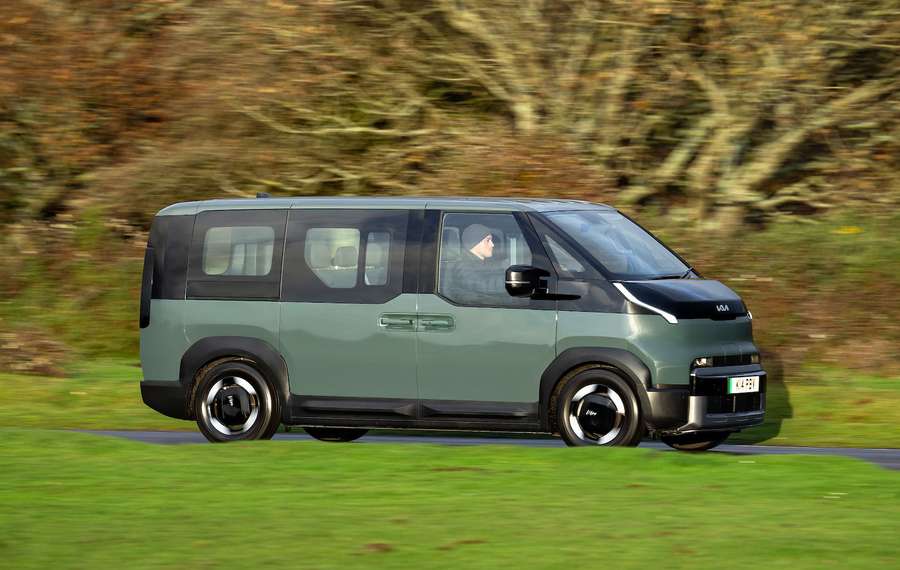 A Kia PV5 with a dark green and black colour scheme driving on a road, set against a blurred, leafy background.