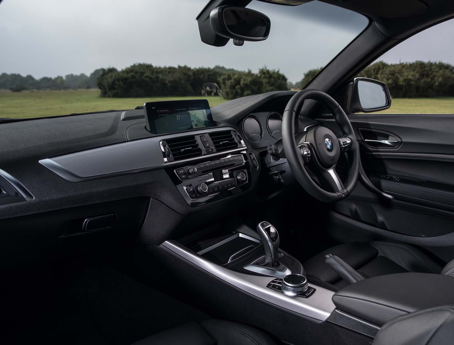 BMW 2 Series interior with a sleek dashboard, steering wheel, infotainment screen, and gear shift. A grassy landscape is visible through the window.