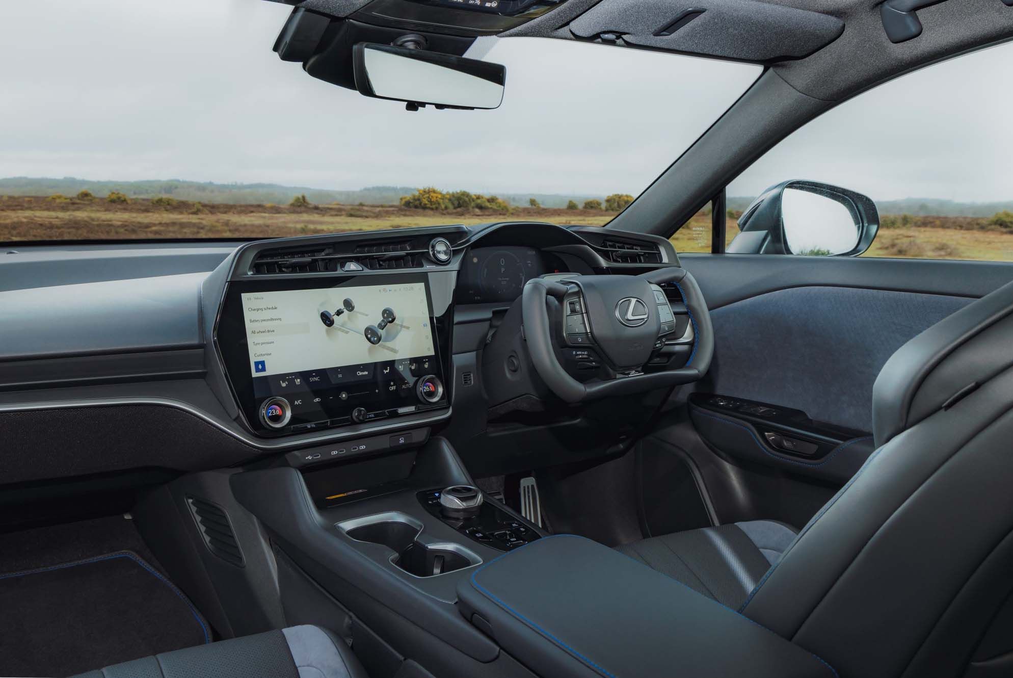 Interior of a Lexus RZ featuring a steering wheel with a logo, a large central touchscreen display, and leather seats, overlooking a scenic landscape.