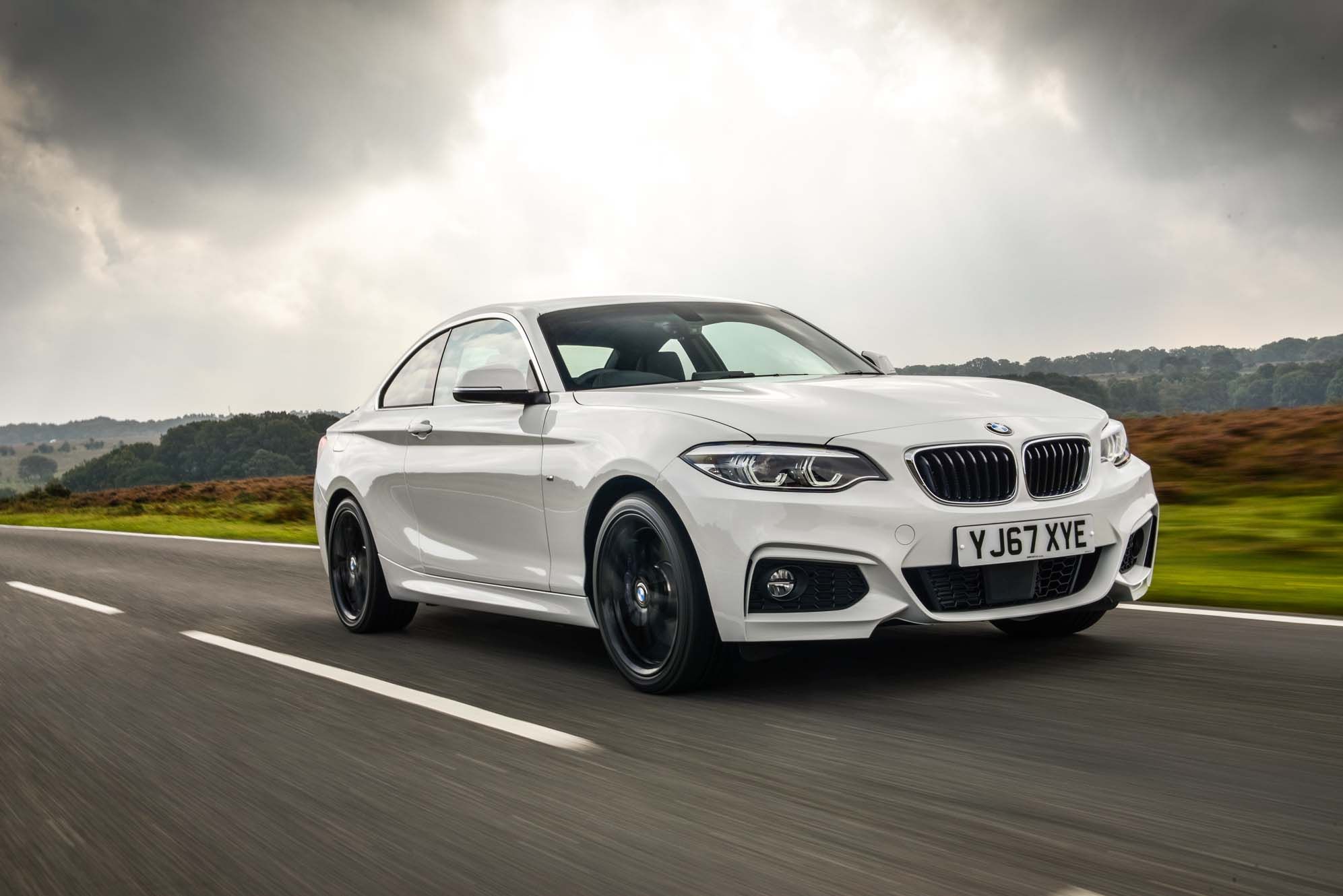 White BMW 2 Series speeding on a scenic country road under a cloudy sky.