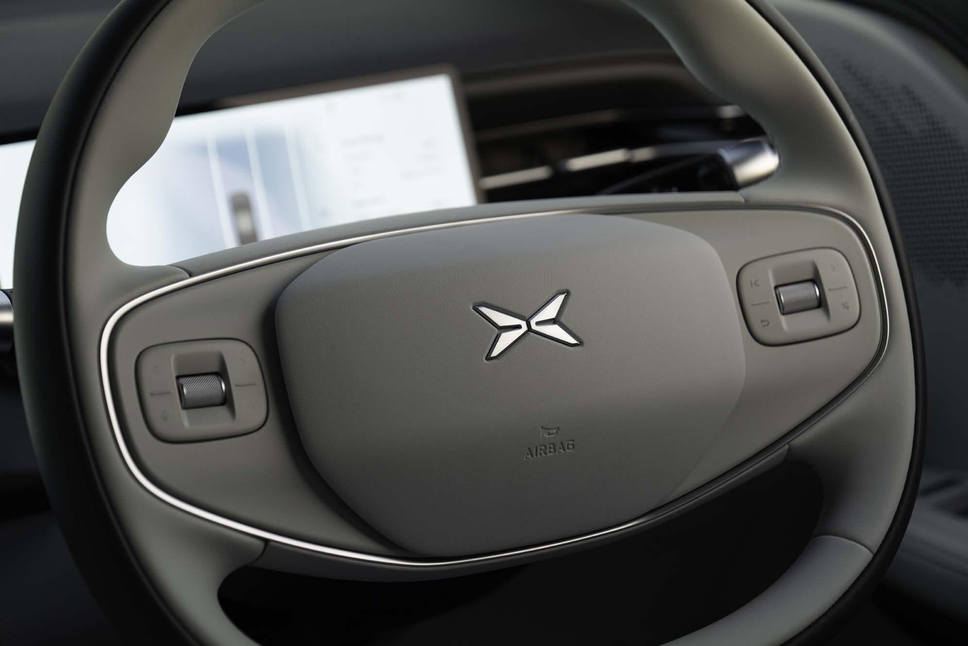 Close-up of an XPENG G6 steering wheel with modern controls and a stylised logo at the centre, in front of a blurred dashboard screen.
