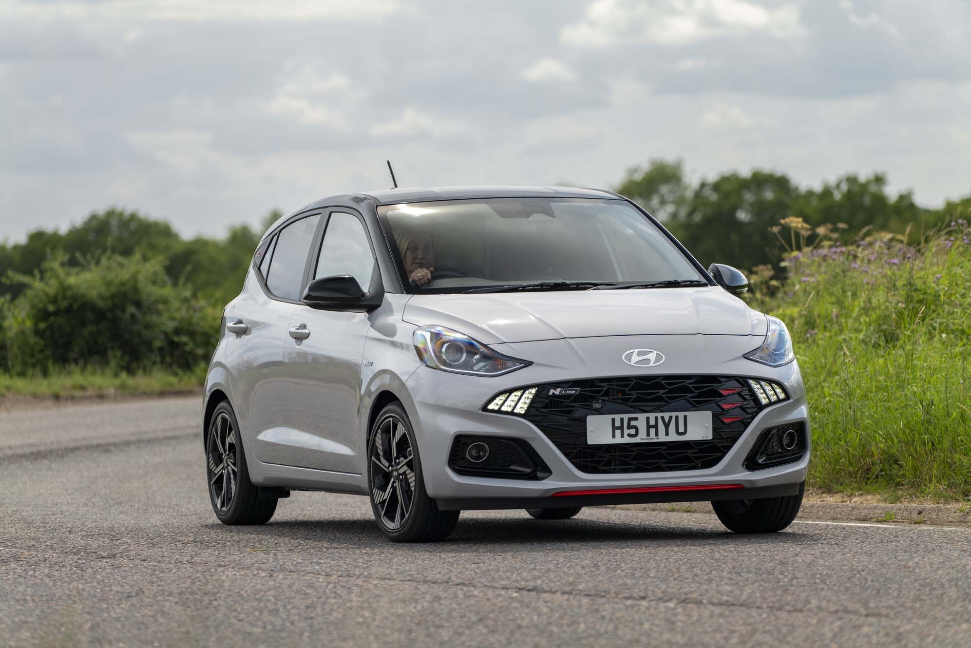Silver Hyundai i10 with sporty design, driving on a road, surrounded by greenery and cloudy sky in the background.