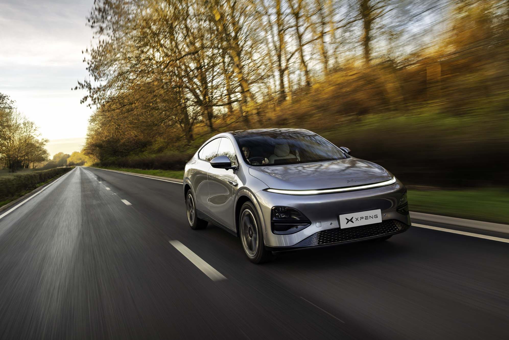 Silver XPENG G6 driving on a scenic road lined with autumn trees and blurred background, under a clear sky.