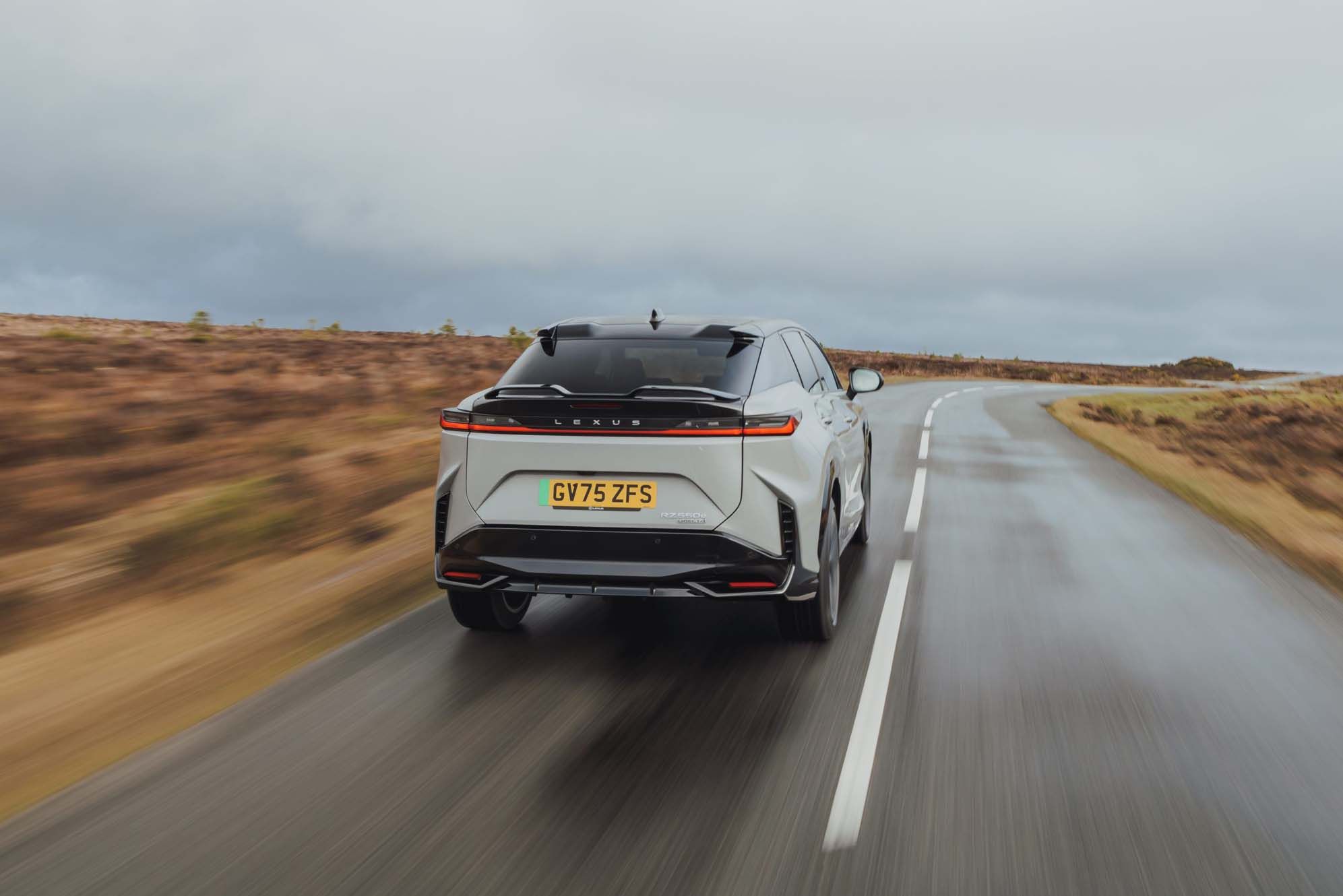 A silver Lexus RZ with a "GV75 ZFS" number plate drives on a wet, winding rural road under a cloudy sky.