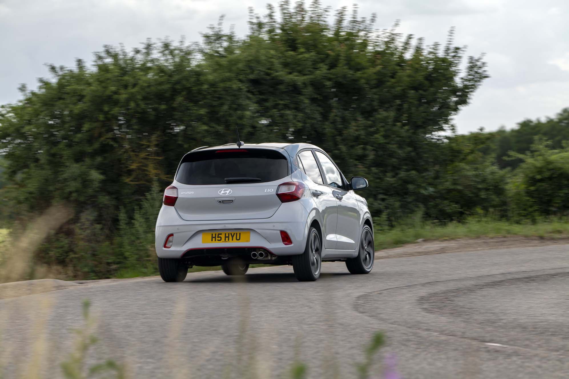 A silver Hyundai i10 driving on a curved road with greenery in the background under a cloudy sky.