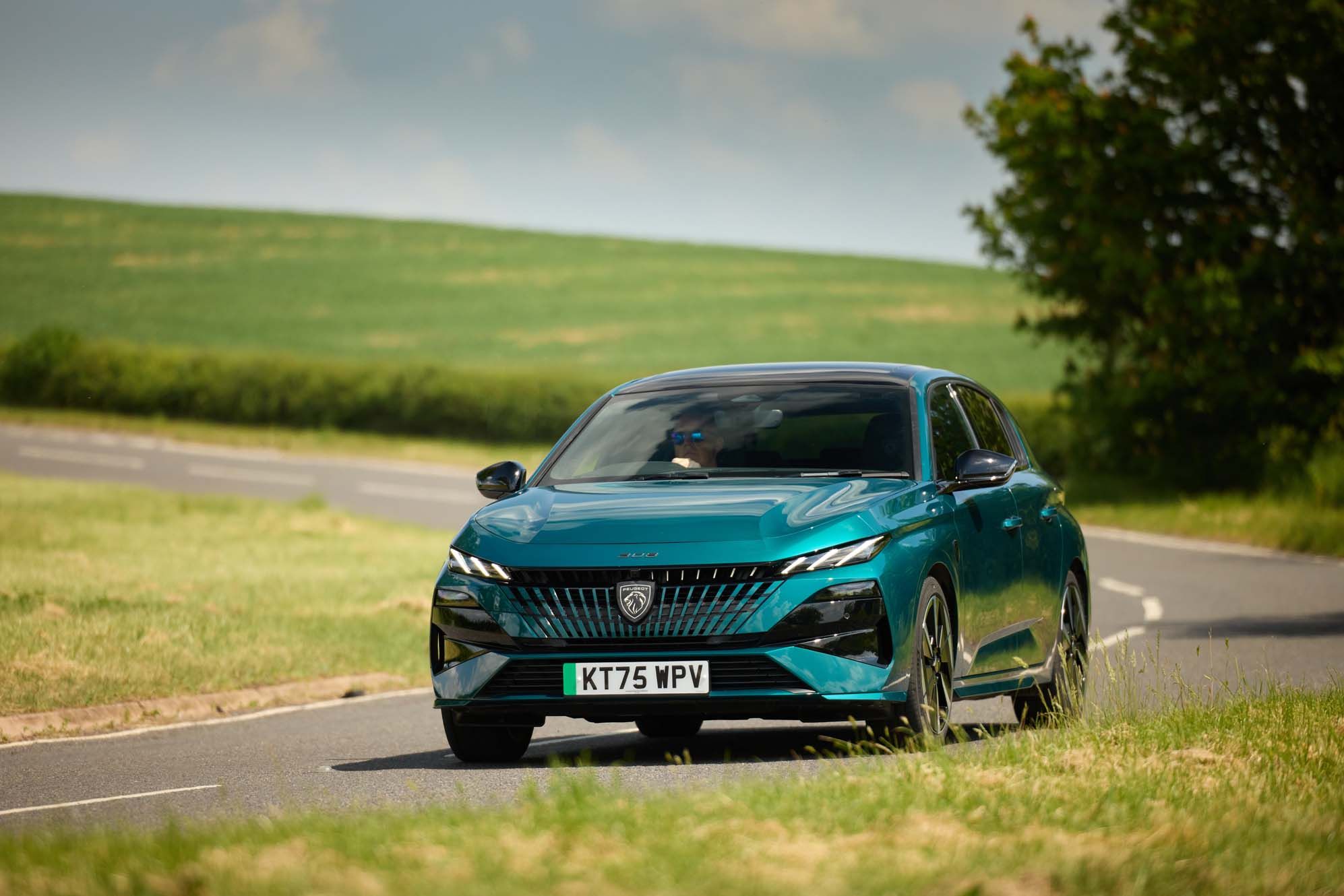 A Peugeot E-308 driving on a rural road surrounded by green fields and trees under a clear blue sky.