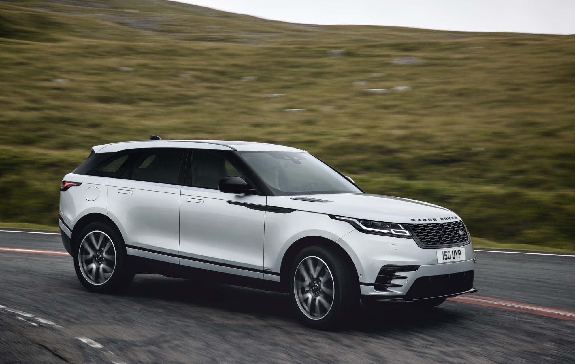 A white Range Rover Velar drives on a curved road with grassy hills in the background.