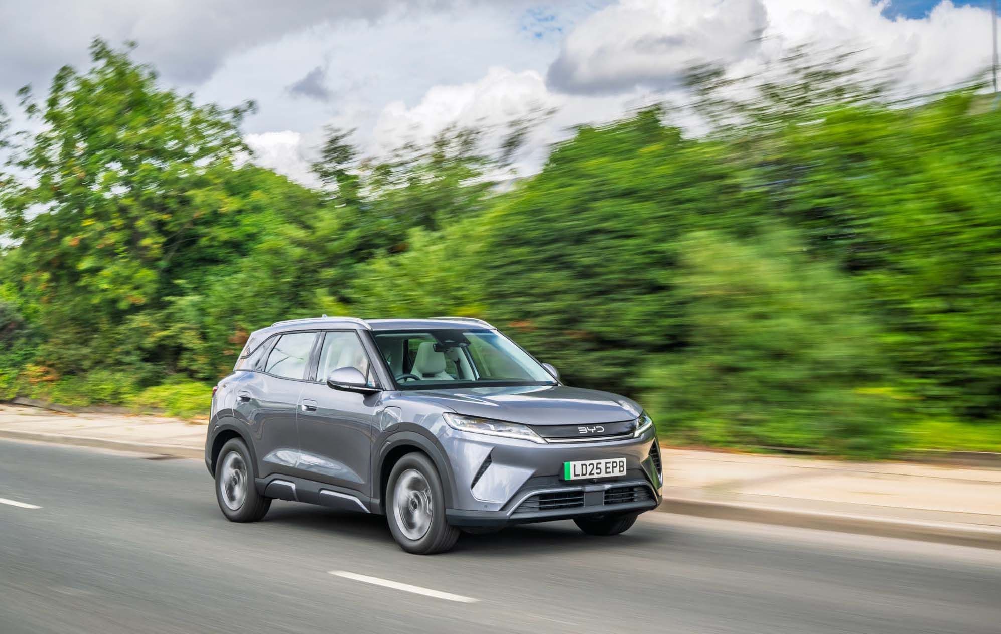 A grey BYD ATTO 2 drives swiftly on a road with blurred greenery in the background under a cloudy sky.