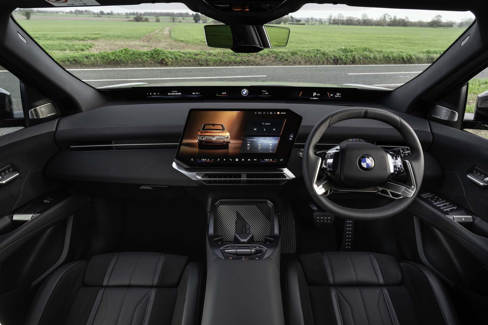 Interior of a BMW iX3 with a high-tech dashboard, large touchscreen display, steering wheel, and leather seats, overlooking a grassy landscape.
