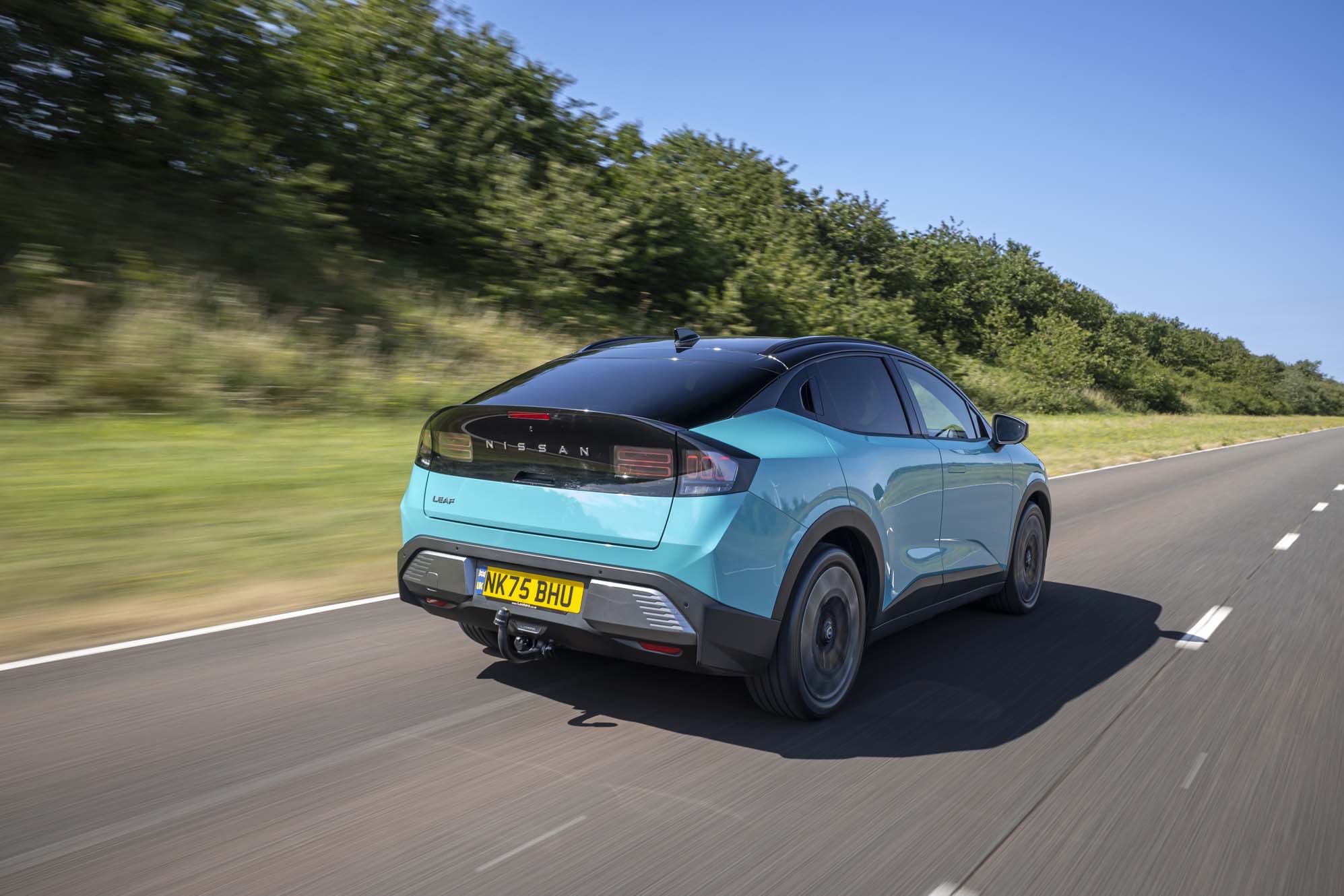 A turquoise Nissan LEAF driving on a clear highway with trees and blue sky in the background.