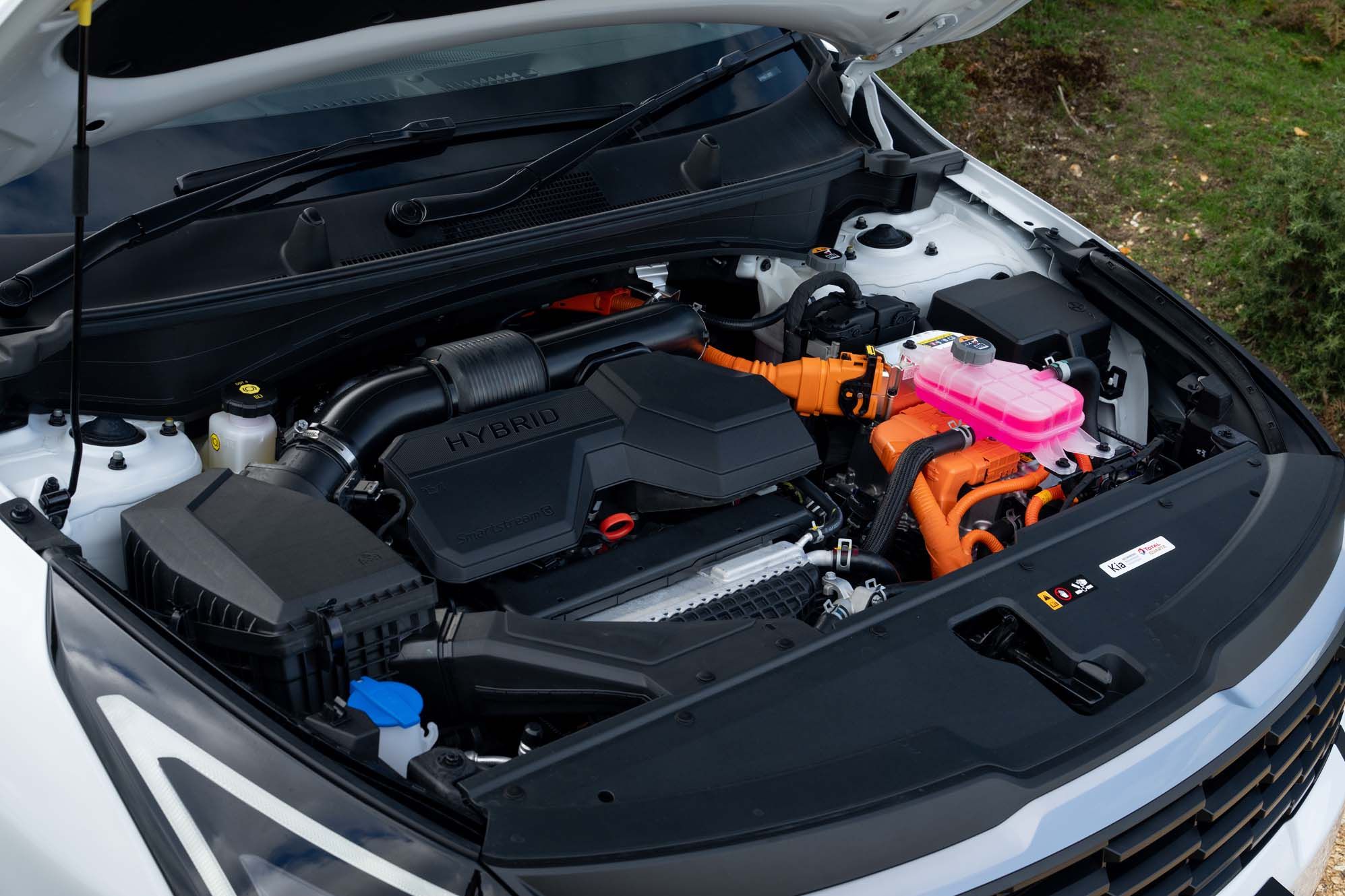Close-up of a Kia Sportage's hybrid engine with orange and pink cables, set within an open bonnet, showcasing modern engineering details.