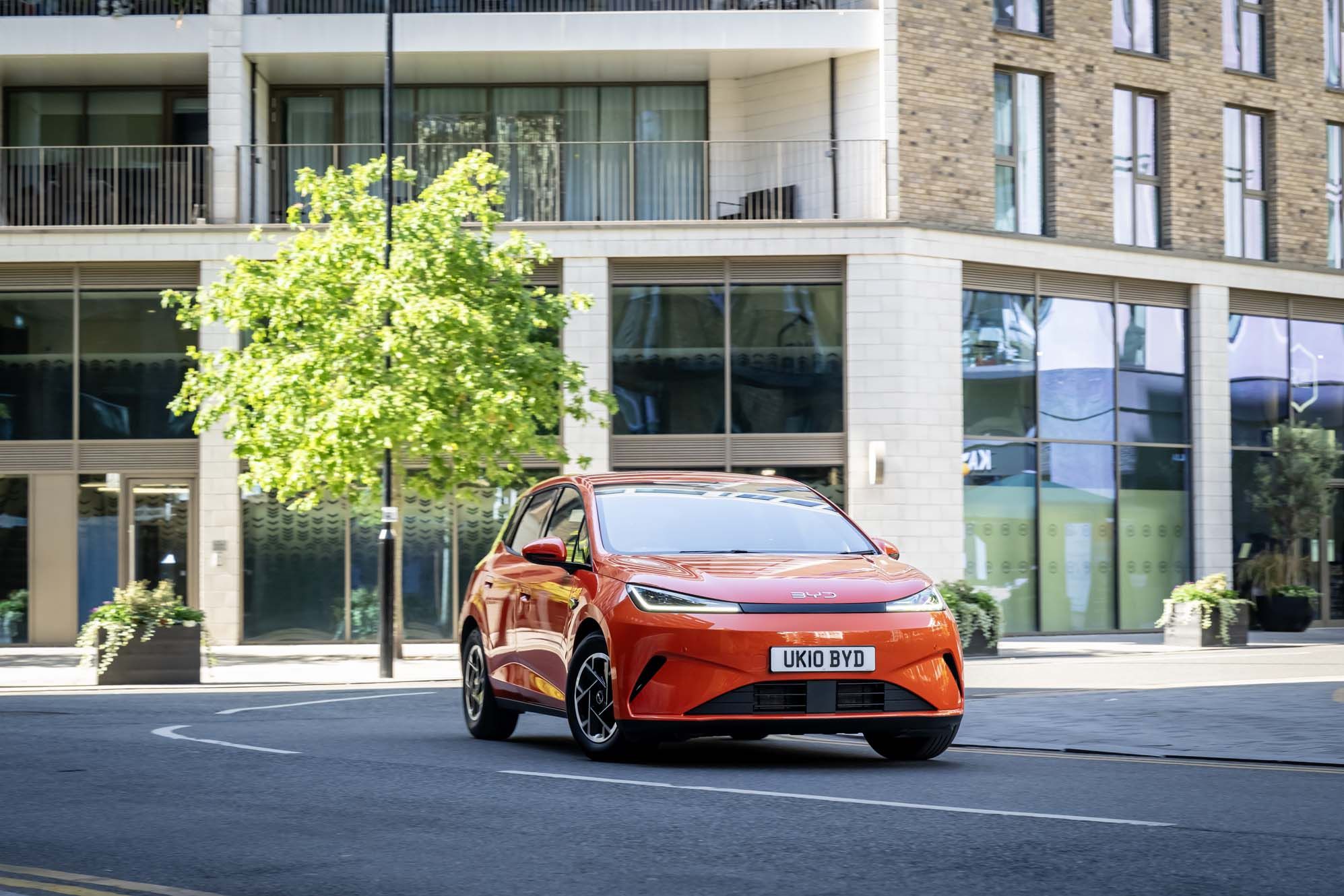 Orange BYD DOLPHIN G DM-i driving on a city street, flanked by modern buildings and a small tree, under clear skies.