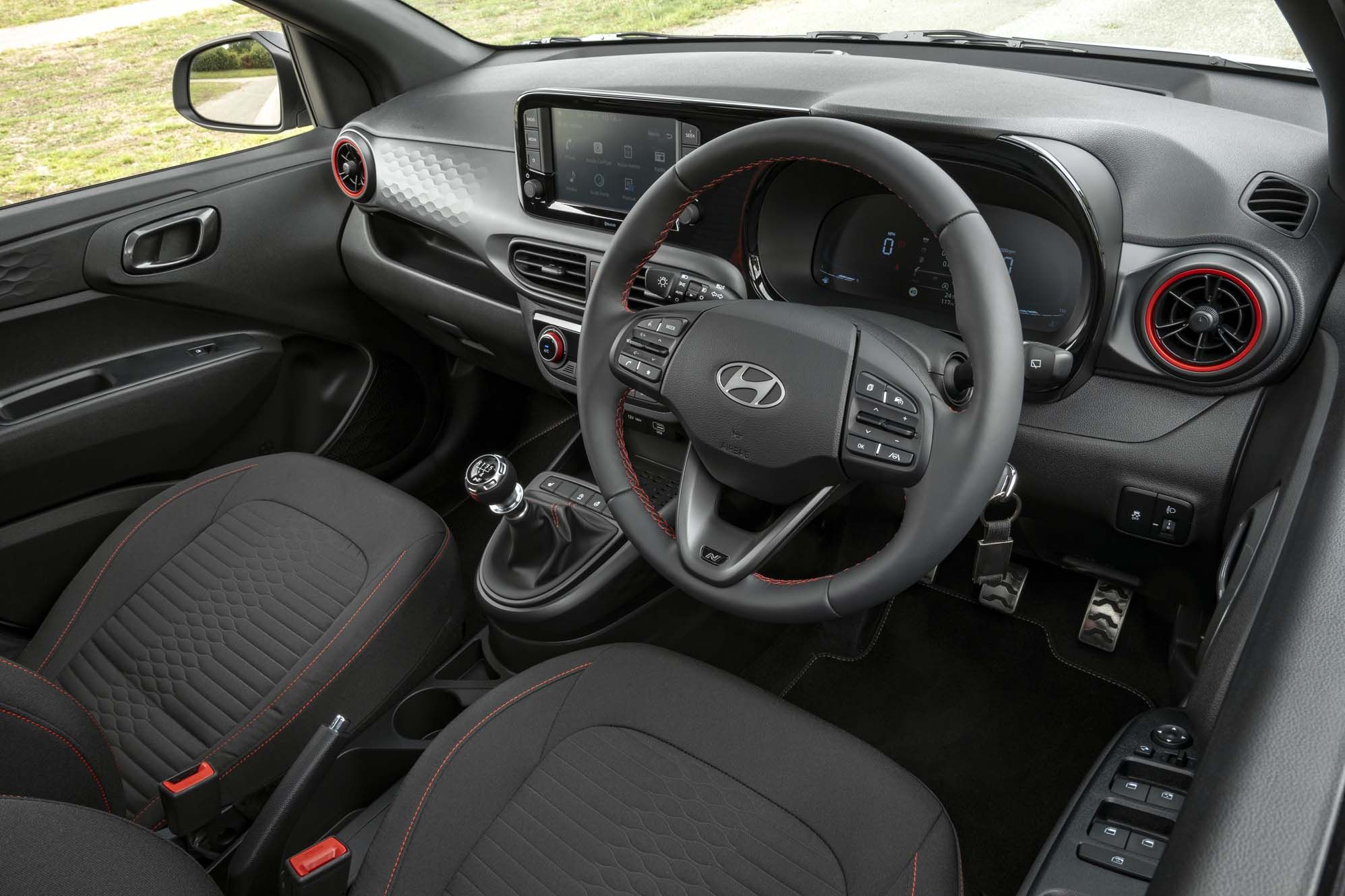 Interior of a Hyundai i10 featuring a sleek dashboard, touchscreen display, manual gear shift, and black seats with red stitching accents.