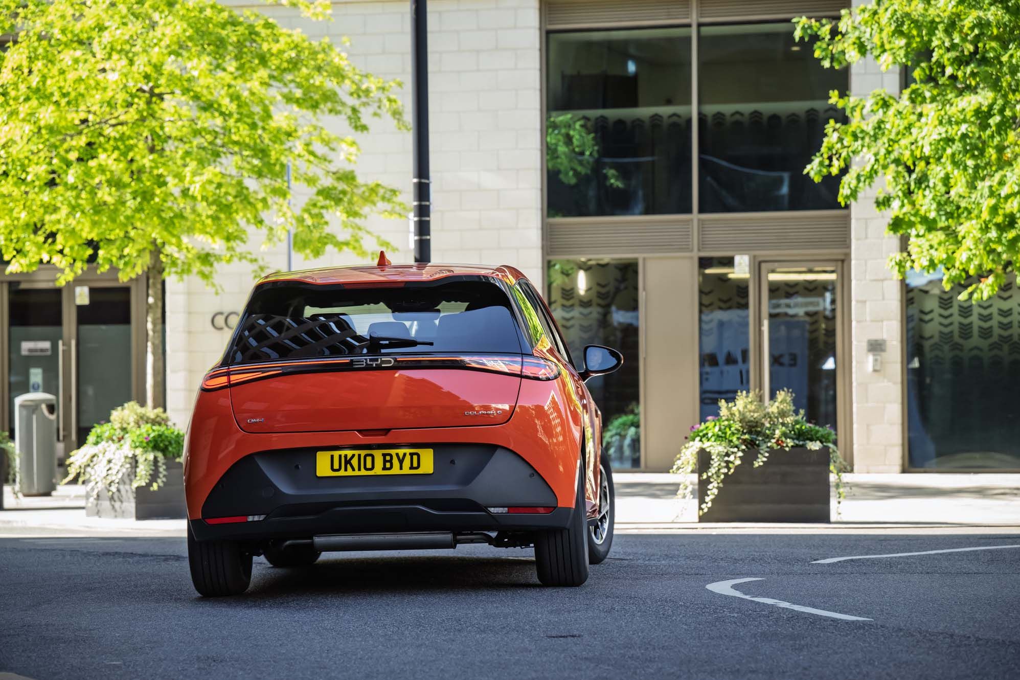 Orange BYD DOLPHIN G DM-i with UK number plate driving on a street, with a modern building and greenery in the background.