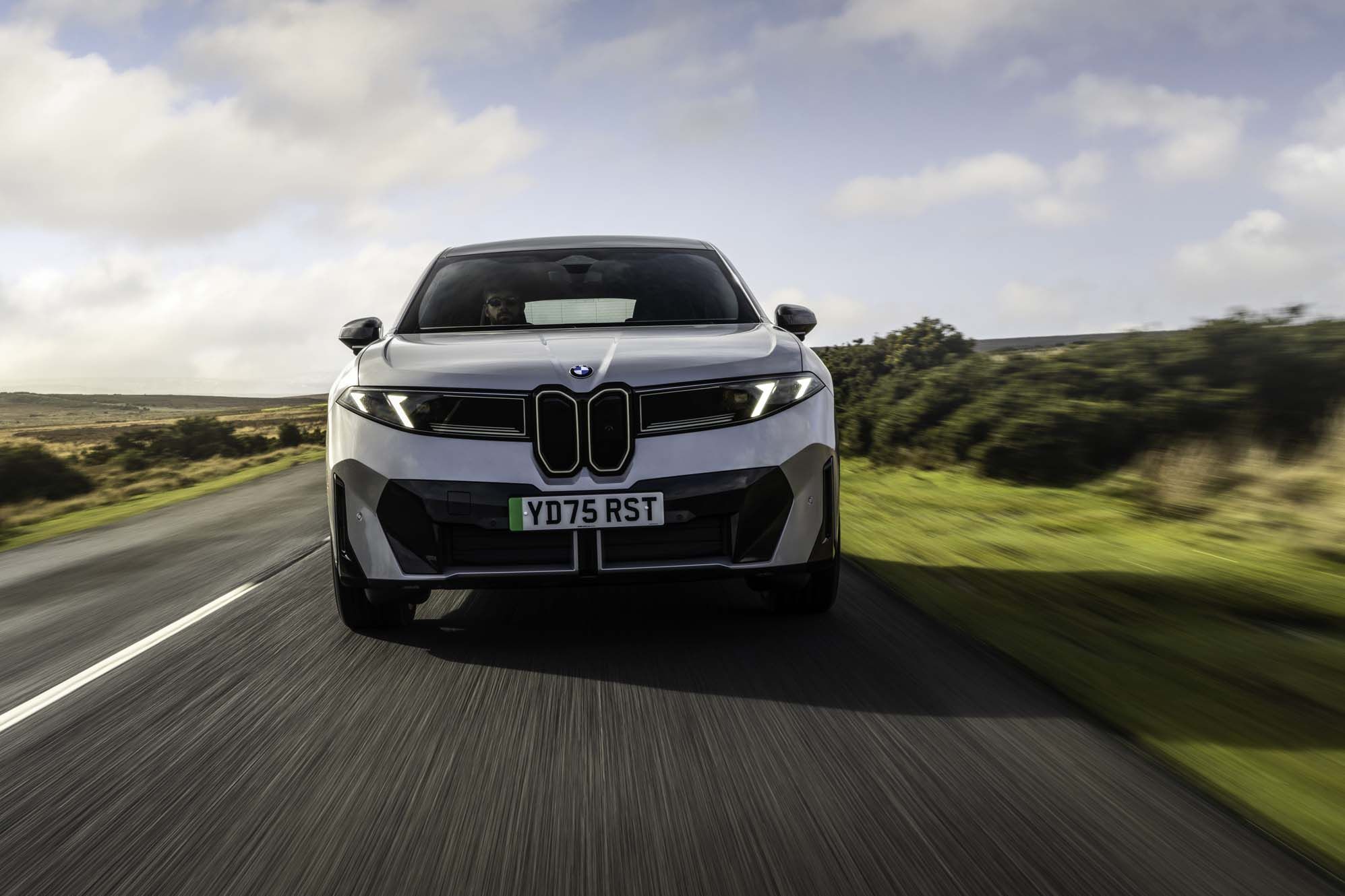 A silver BMW iX3 on an open road, surrounded by countryside, under a partly cloudy sky.