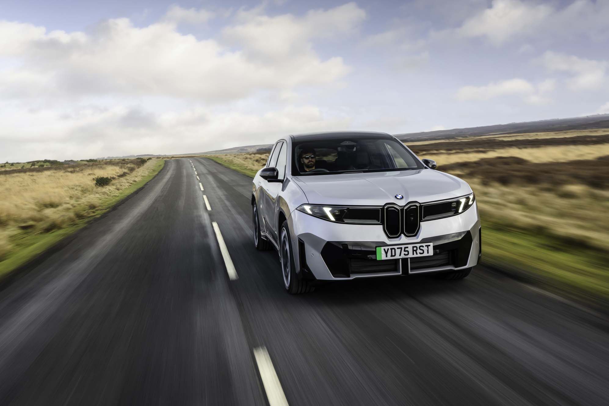 A silver BMW iX3 drives on a deserted rural road with grassy fields and a cloudy sky in the background.
