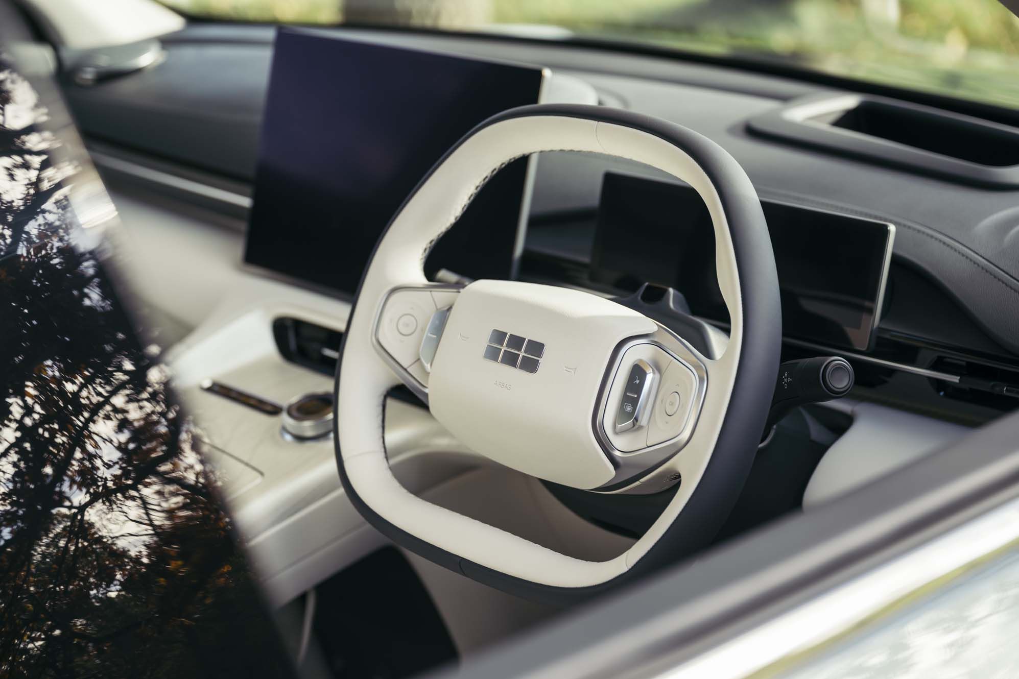 Geely EX5 interior with a sleek steering wheel featuring control buttons, digital dashboard screens, and reflected trees on the windshield.